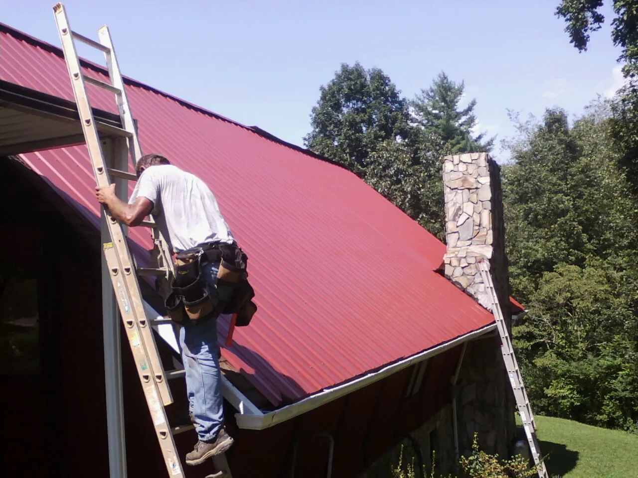 Roofer on a ladder beside a red metal roof and stone chimney — Soffit & Fascia Repair by Roofing Brothers in Brooktondale